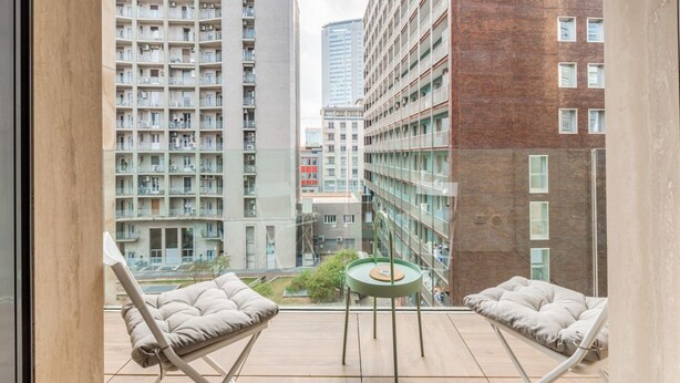 Un balcón con dos sillas acolchadas y una pequeña mesa, con vistas a un paisaje urbano de rascacielos.