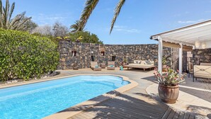 A swimming pool with a wooden deck, stone walls, and outdoor seating. - House with heated swimming pool, solarium, large garden, high-speed WIFI access. (Lajares)