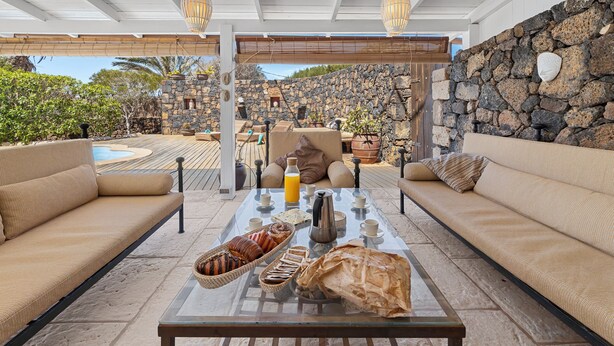 A sunlit outdoor seating area with a stone wall, a pool, and a breakfast setup on a glass table. - House with heated swimming pool, solarium, large garden, high-speed WIFI access. (Lajares)