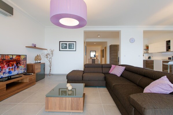A modern living room with a sectional sofa, a glass coffee table, and a television on a wooden stand. There is a large purple pendant light, a framed picture on the wall, and a view into the kitchen area.