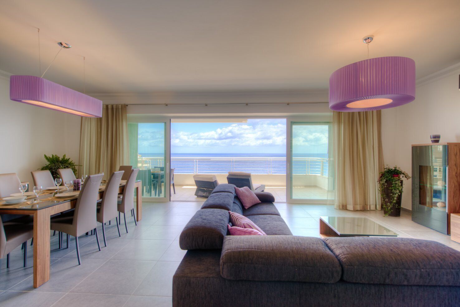 Sala de estar moderna com sofá, mesa de jantar e cadeiras, varanda com vista para o mar e uma janela grande.