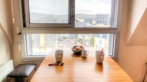 A room with a wooden table, two mugs, a bowl of pastries, and a view of a town through large windows. - Nice apartment for 2 people with WIFI and TV (Trouville-sur-mer)