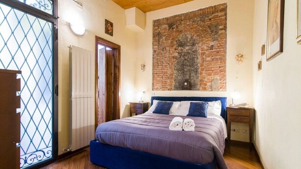 A bedroom with a bed, wooden floor, a brick wall, and a window with a metal grill.
