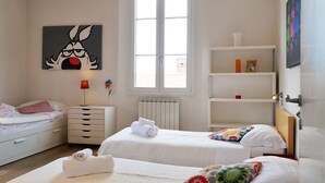 Una camera d'albergo con due letti, un termosifone, una finestra, uno scaffale con oggetti decorativi e un quadro appeso al muro.