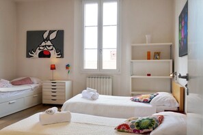Una habitación de hotel con dos camas, un radiador, una ventana, una estantería con adornos y un cuadro en la pared.