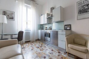 A modern kitchen with white cabinets, a gas stove, and a built-in oven. There is a dining table with chairs, a sofa, and a radiator.