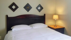 Una cama bien hecha con sábanas blancas, un cabecero de madera oscura y una mesita de noche con una lámpara. Hay tres espejos cuadrados en la pared, sobre la cama.