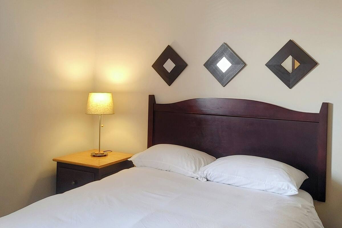 Ein ordentlich bezogenes Bett mit weißen Leinen, ein Holzkopfschild und ein Nachttisch mit Lampe. Darüber an der Wand hängen drei Dekoartikel.