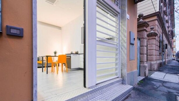 A building entrance with a sliding door, a small outdoor seating area with chairs, and a paved sidewalk.
