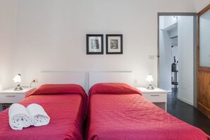 A hotel room with two beds, a nightstand with a lamp, a wall-mounted picture, and a door leading to another room.