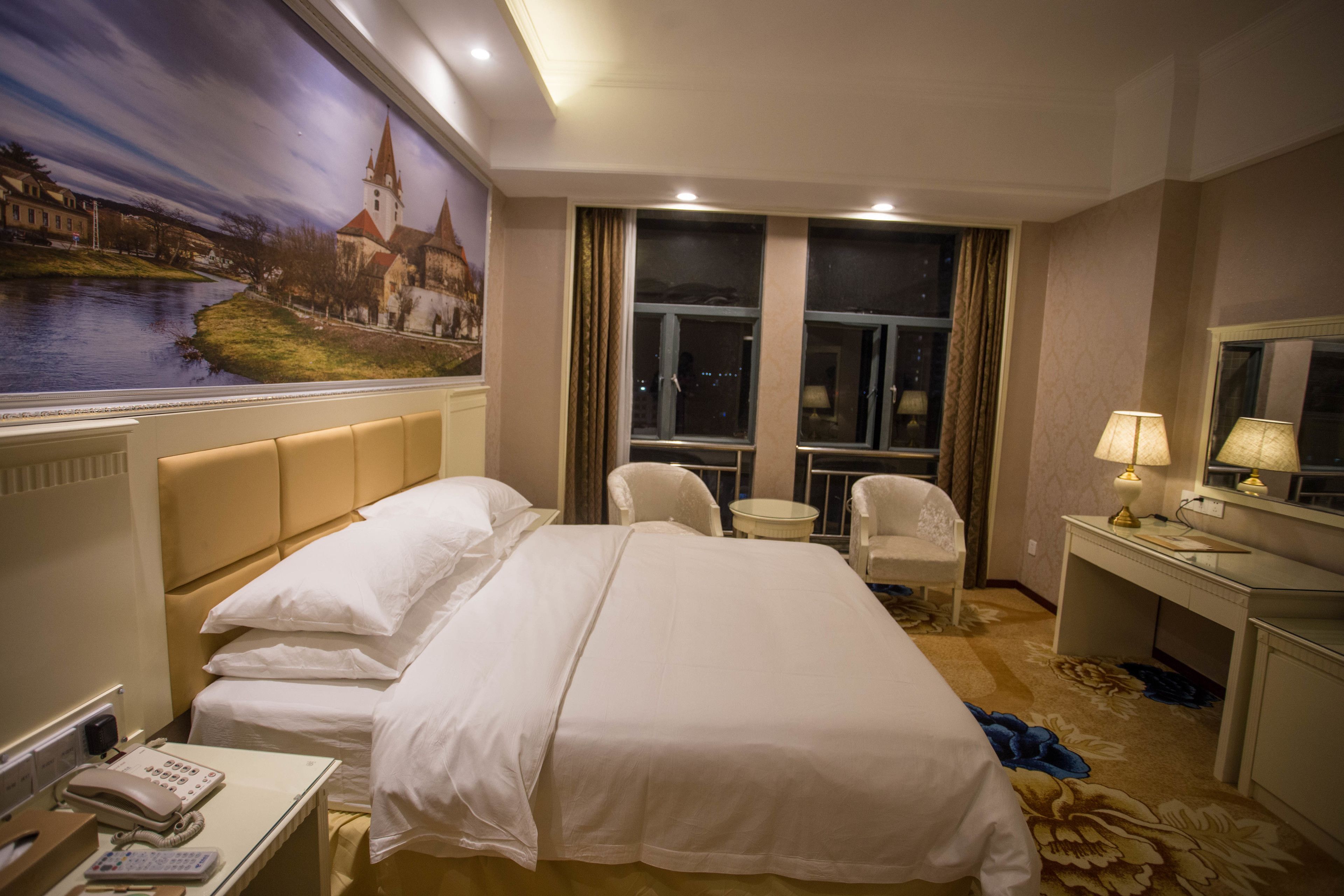 A hotel room with a large bed, a desk, a chair, a small table, a telephone, and a painting on the wall.