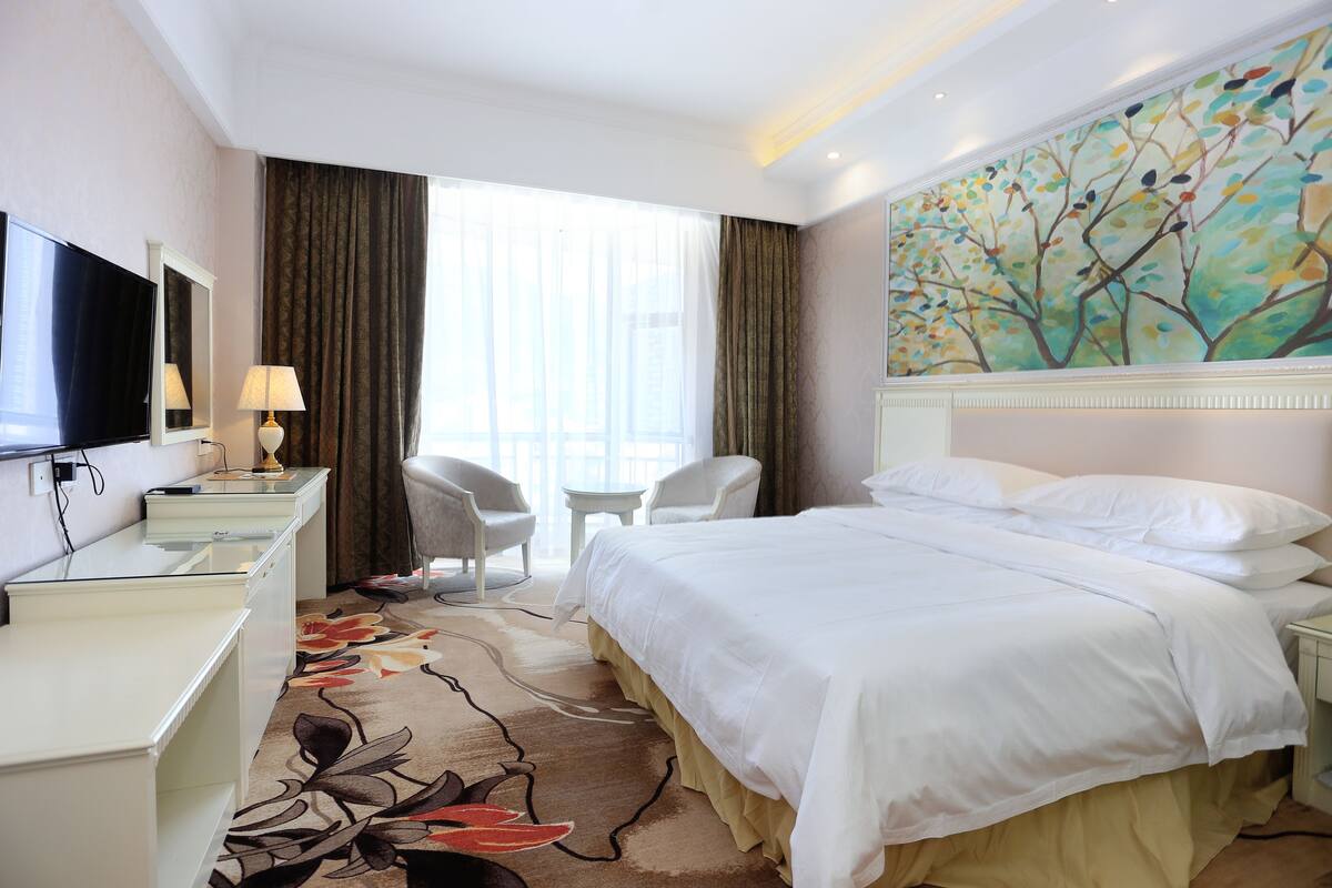Een hotelkamer met een bed, een bureau met een televisie, een stoel, een klein tafeltje en een schilderij aan de muur.