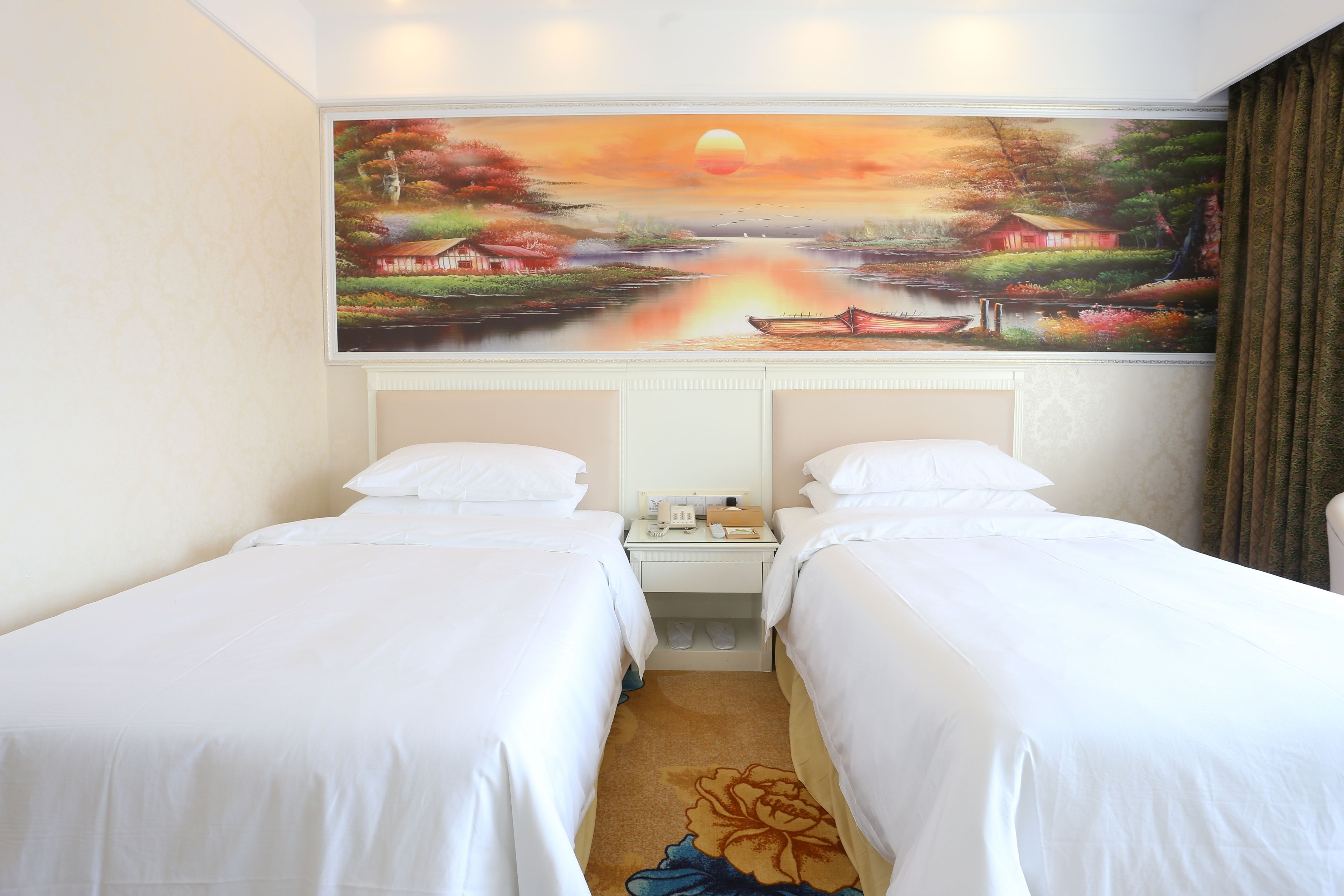 A hotel room with two beds, a painting of a sunset landscape on the wall, and a carpet with a floral design.