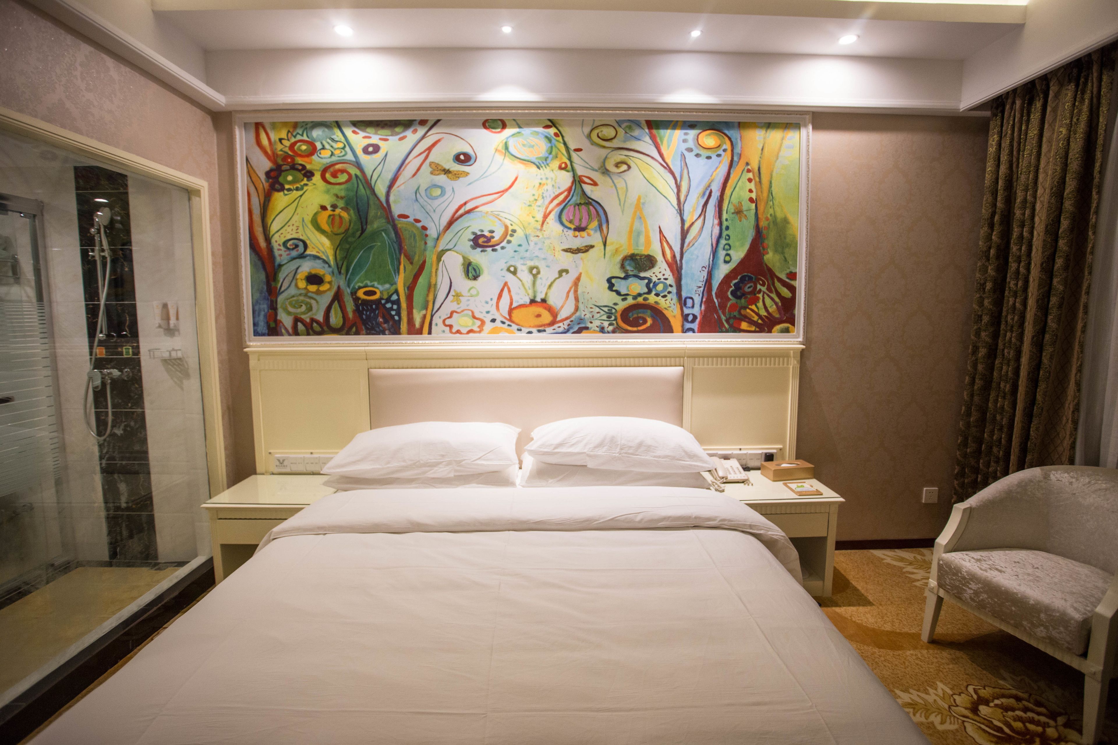Habitación de hotel con una cama grande, una mesita de noche con teléfono, una silla y un cuadro abstracto de colores en la pared.