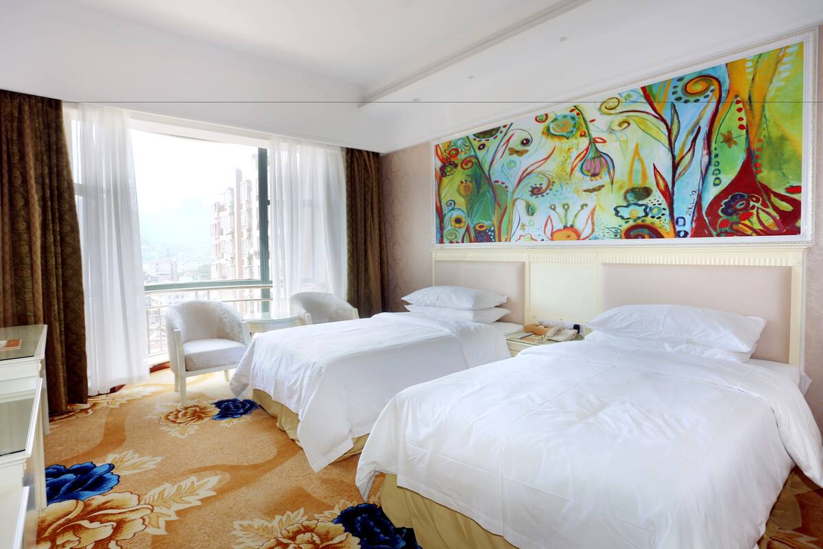 Een hotelkamer met twee bedden, een stoel, een bureau en een groot, kleurrijk schilderij aan de muur.