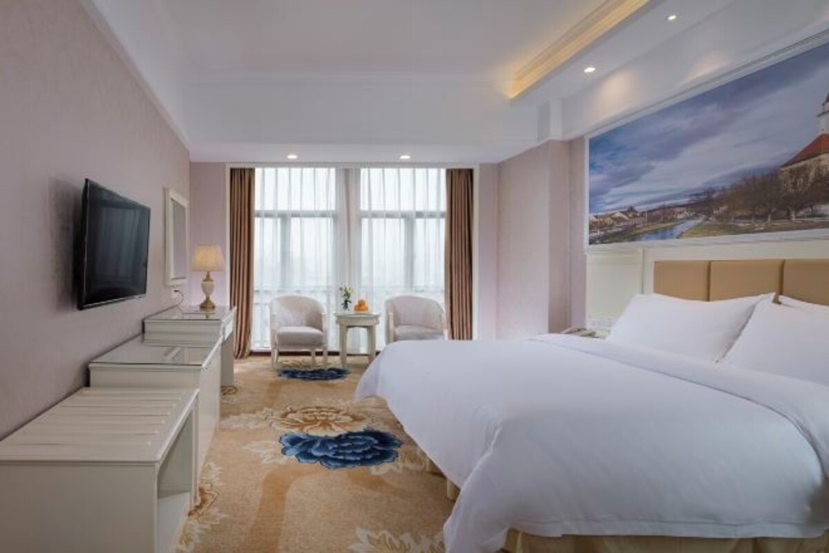 Une chambre d’hôtel avec un grand lit, un téléviseur à écran plat fixé au mur, une petite table avec une lampe et un tableau représentant un paysage accroché au mur.