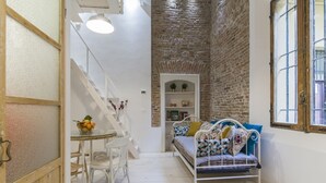 A narrow room with a white sofa, a small table with chairs, a staircase, and a brick wall.