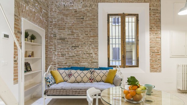 A room with a brick wall, a white sofa with patterned cushions, a glass table with a bowl of fruit, and a window with a metal grill.
