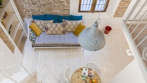 A loft-style room with a high ceiling, exposed brick walls, a wooden floor, a sofa with patterned cushions, a dining table with chairs, and a hanging light fixture.