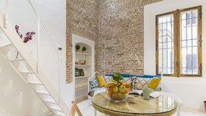 A room with a brick wall, a white staircase, a glass table with a fruit bowl, a white sofa with cushions, and a window with wooden frames.