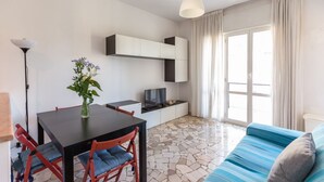 A modern living room with a dining table, chairs, a sofa, a TV, and a large window with curtains. - GuestHost - NEW - Forlanini apartment for 4! (milano)