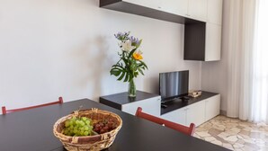 A modern dining area with a black table, red chairs, a basket of fruit, a vase with flowers, and a TV on a cabinet. - GuestHost - NEW - Forlanini apartment for 4! (milano)