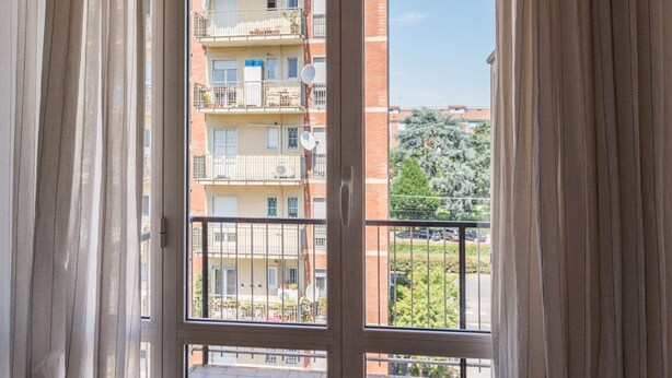 A view through a window with curtains, revealing a balcony with a metal railing, a multi-story building with balconies, and a clear sky. - GuestHost - NEW - Forlanini apartment for 4! (milano)