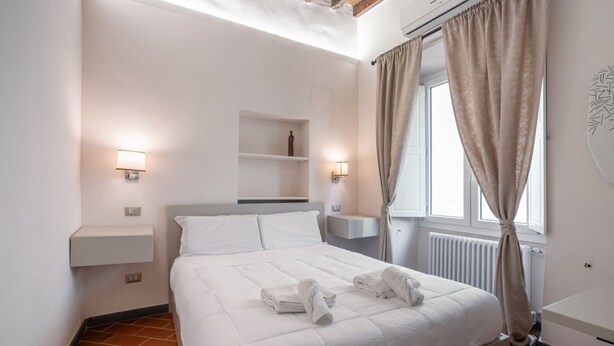 Une chambre d’hôtel avec un lit, des lampes de chevet, une étagère et une fenêtre avec des rideaux.