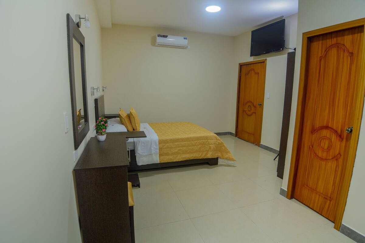 Habitación de hotel con cama, escritorio con silla, televisor montado en la pared y puerta de madera.