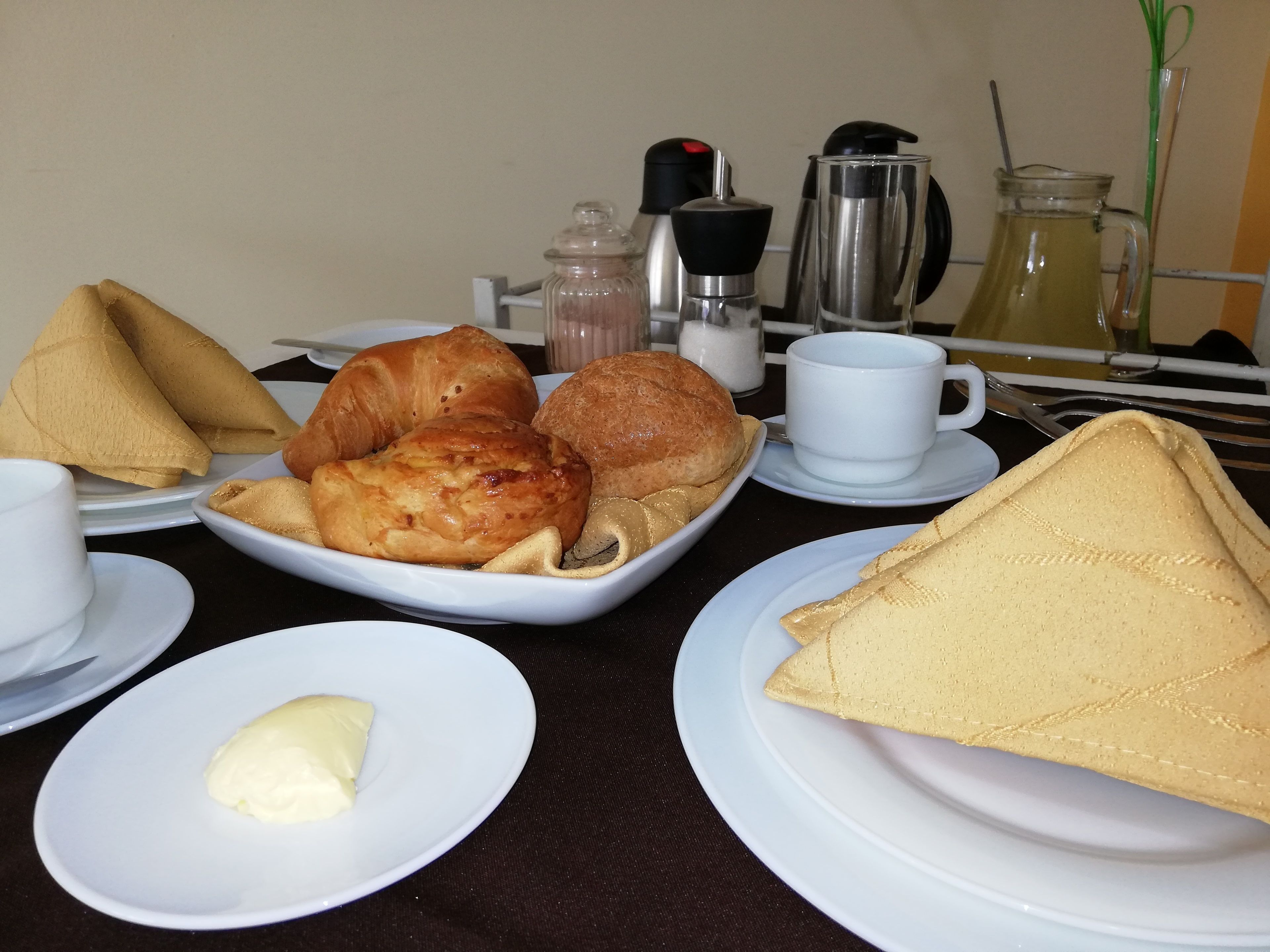 Una mesa de desayuno con croissants, un sándwich de croissant, un sándwich de croissant con una loncha de queso, un plato pequeño con mantequilla, una taza y una jarrón.