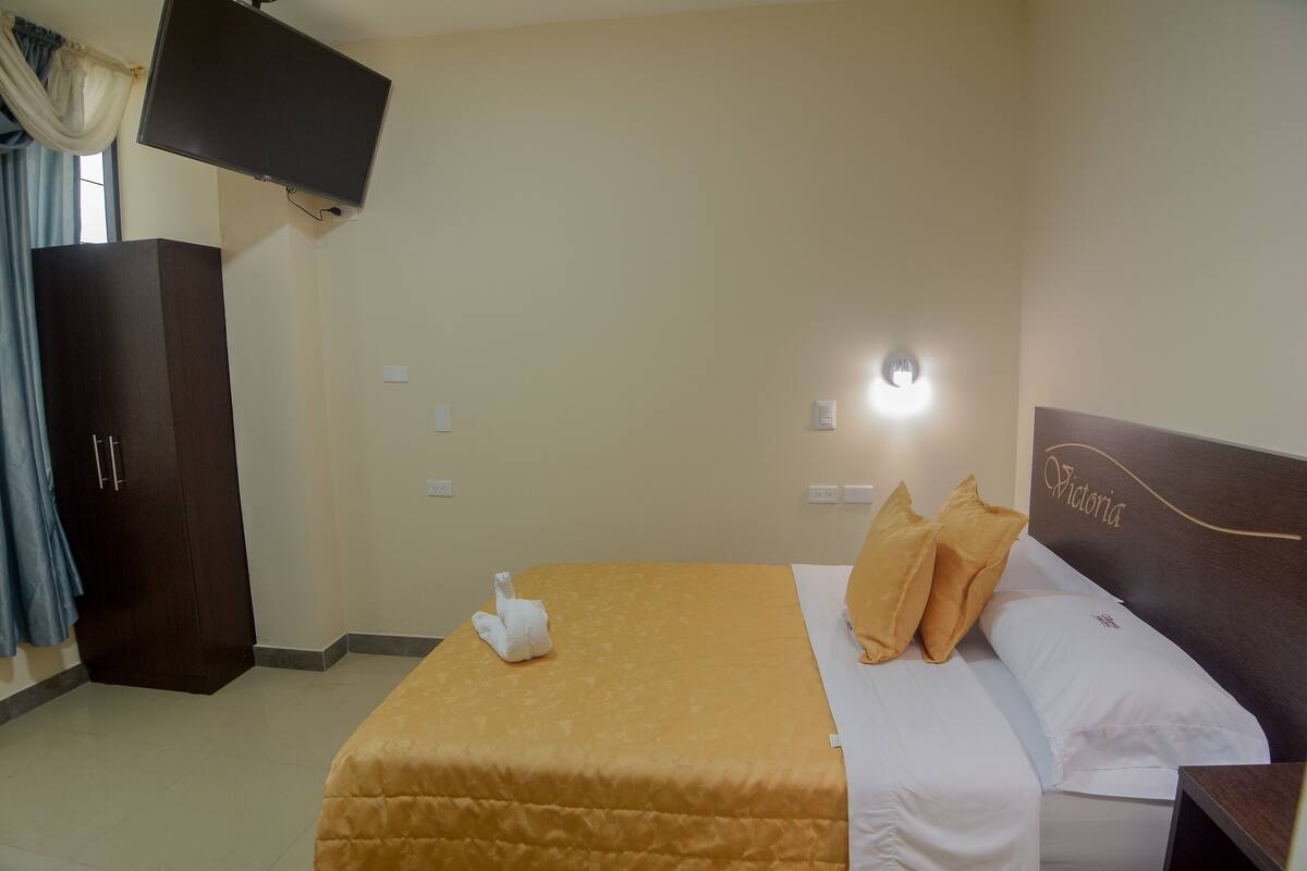 Una habitación de hotel con cama, televisor montado en la pared, armario y mesita de noche.
