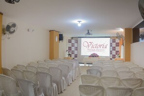 A conference room with rows of white chairs, a projector screen displaying "Victoria Suites Hotel", and a stage with a table and flowers. - Victoria Suites Hostal (Santo Domingo de los Colorados)