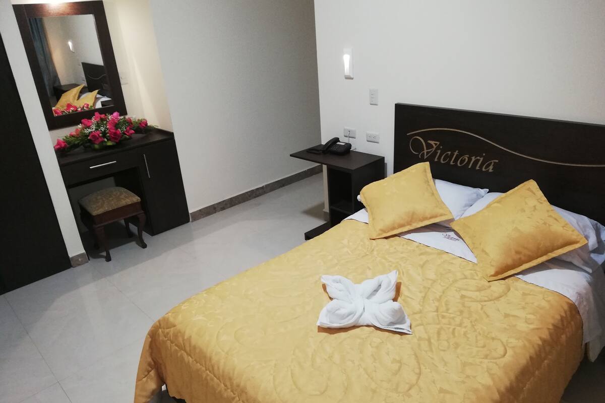 Habitación de hotel con cama, mesita de noche, escritorio y silla, espejo y una placa con el nombre en la pared.