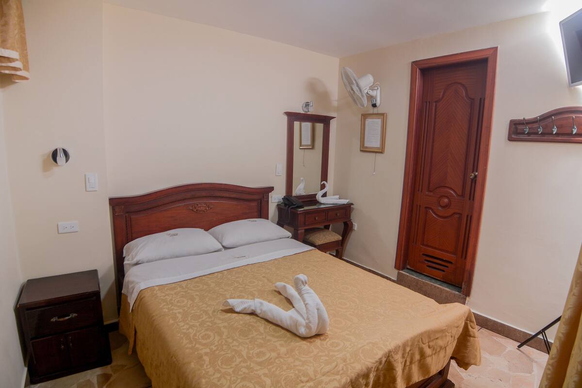 Habitación de hotel con cama, mesita de noche, cómoda, espejo, puerta y ventilador de pared.