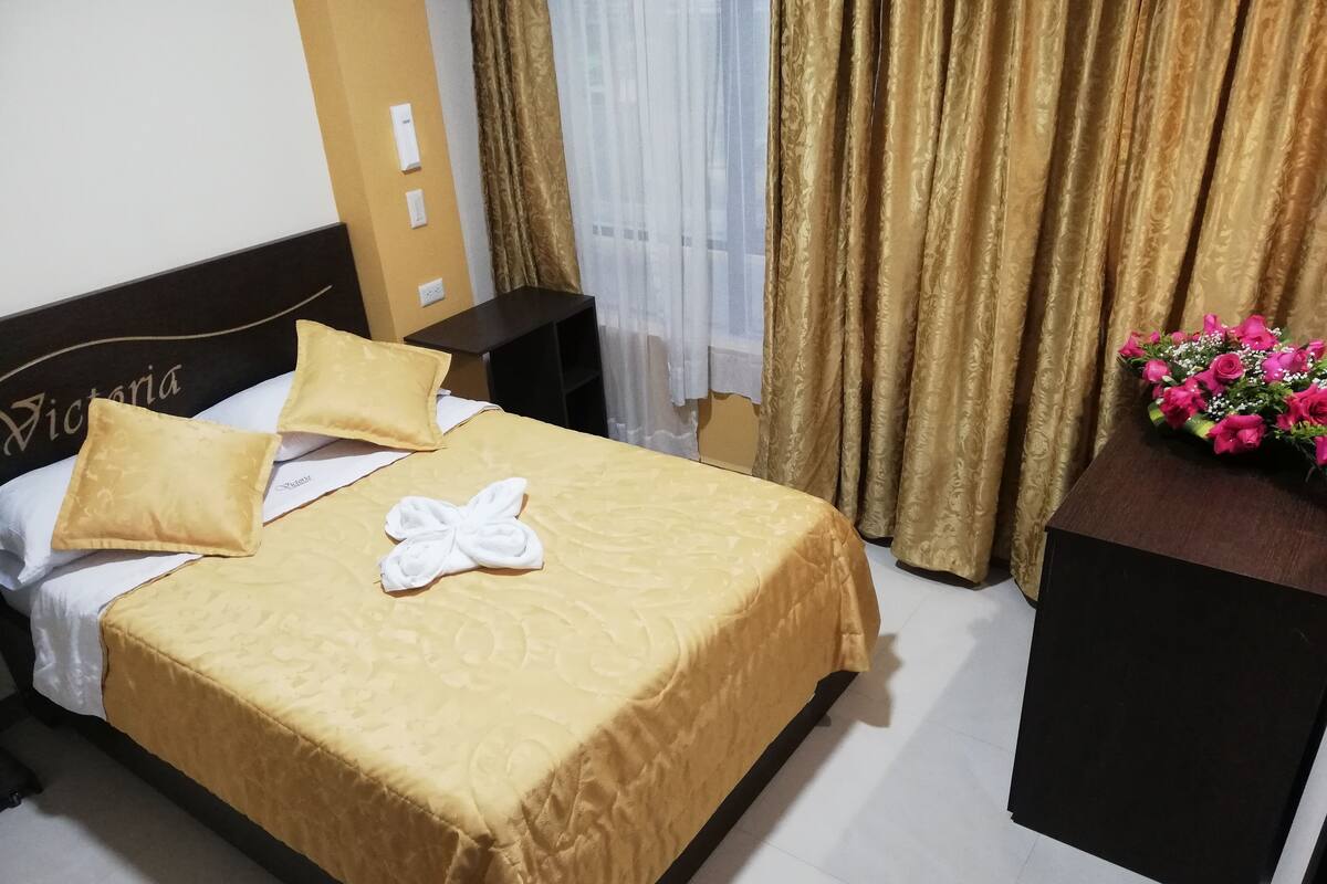 Una cama bien hecha con una colcha amarilla, una almohada blanca y una toalla doblada en forma de lazo. Junto a la cama hay una mesita de noche de madera oscura con un pequeño ramo de flores rosas encima. La habitación tiene una pared color beige, una señal con la inscripción 'Victoria', cortinas doradas y una persiana blanca.