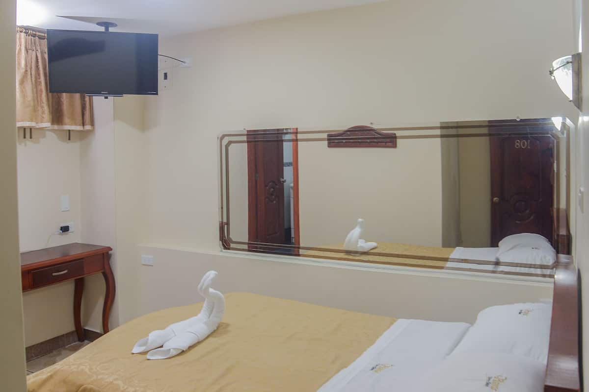 Una habitación de hotel con una cama, televisión en la pared, un espejo y un escritorio de madera.