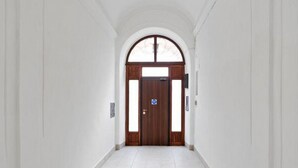 Un couloir menant à une porte en bois, sur laquelle est apposée une plaque, avec des murs blancs et un sol carrelé.