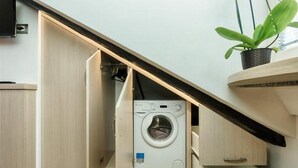 A compact laundry room with a washing machine, a wooden cabinet, and a potted plant. - GuestHost - Cozy and stylish studio situated in the centre of Turin in a beautiful historical building with elevator. It is the ideal solution for both short business or leisure stays: the area has a good infrastructure and is well served by lots of faci (Turin)