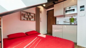 A bedroom with a red bedspread, a kitchen with white cabinets, and a wooden ceiling beam. - GuestHost - Cozy and stylish studio situated in the centre of Turin in a beautiful historical building with elevator. It is the ideal solution for both short business or leisure stays: the area has a good infrastructure and is well served by lots of faci (Turin)