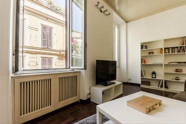 A modern living room with a large window, a flat-screen TV, a bookshelf, and a white coffee table.