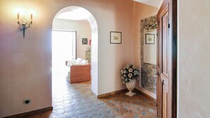 Un pasillo con suelo de baldosas, un aire acondicionado de pared, un aplique, un arco, una cesta de mimbre, un jarrón con flores y una pared de piedra con cuadros enmarcados.