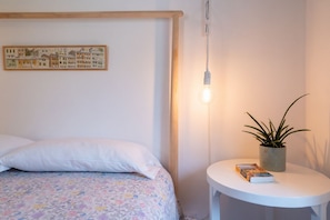 A bedroom with a bed, a bedside table with a plant and a book, a wall-mounted light, and a framed picture.