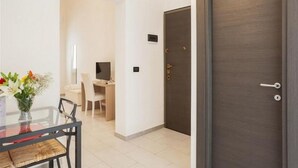 Een moderne hotelkamer met een glazen eettafel, een bloemenvaas, een bureau en een donkere houten deur.