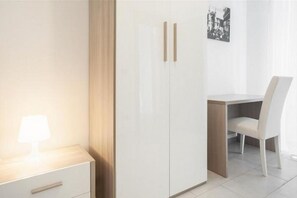 A room with a bed, a bedside table with a lamp, a wardrobe, a desk with a chair, and a framed picture on the wall.