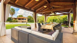 An outdoor covered patio with wicker furniture, a dining table set for six, and a pool in the background. - ClickSardegna Villa Paradiso with swimming pool and exclusive tennis court (Alghero)