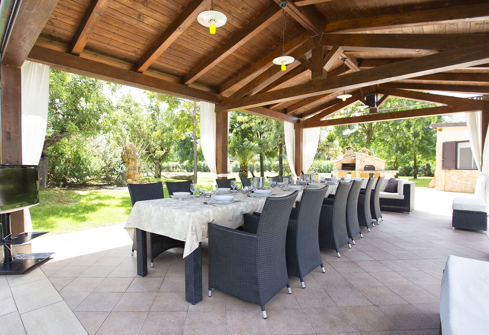 An outdoor covered dining area with a long table set for a meal, surrounded by wicker chairs, under a wooden roof structure.