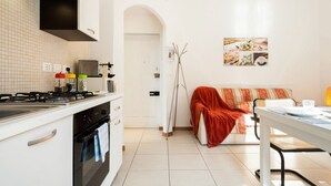 A compact kitchen with a stove, oven, and white cabinetry. A dining area with a table and chairs is visible, and a sofa with a red blanket is in the background. - GuestHost - Casa Panigale Comfortable Apartment x4! NEW (Bologna)