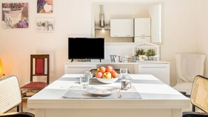 A dining area with a table set for two, a bowl of fruit, a television, and a kitchen in the background. - GuestHost - Casa Panigale Comfortable Apartment x4! NEW (Bologna)