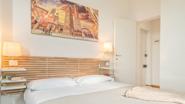 A hotel room with a bed, bedside lamps, a nightstand, a wall-mounted painting, and a door. - GuestHost - Casa Panigale Comfortable Apartment x4! NEW (Bologna)