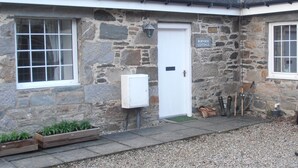 A stone cottage with a white door, a sign reading "Burnside Cottage," and a small garden area with plants and a wooden planter. - Escape to Rural Highlands Stunning Scenery, Sound of the Burn  Licence PK11943F (Aberfeldy)
